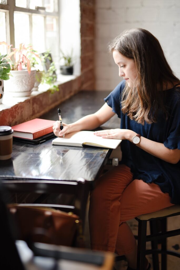 woman sitting at a black table writing in a journal near window