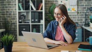 Beautiful woman using laptop and talking on mobile phone in workplace sitting at desk smiling speaking and looking at screen. Communication and job concept.